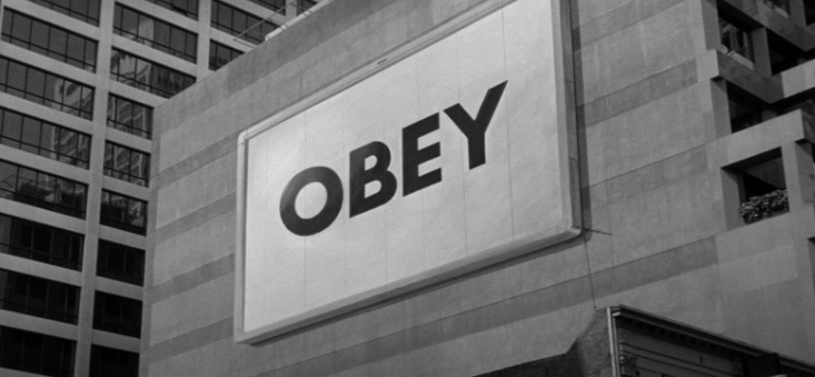 A black-and-white photograph of an urban cityscape featuring a large, illuminated billboard mounted on the side of a modern concrete building, prominently displaying the word "OBEY" in bold, black capital letters on a white background, with tall office buildings visible in the background.