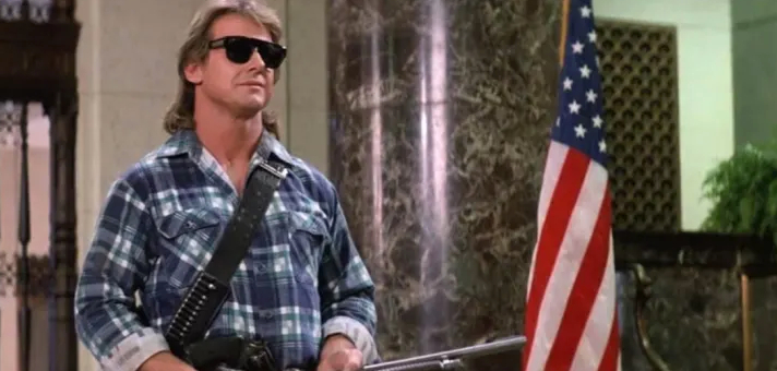 This image shows a scene from the 1988 science fiction film "They Live." The person is wearing dark sunglasses and a blue plaid flannel shirt, positioned near an American flag. The setting appears to be outdoors with some foliage and a light green building or structure in the background. This appears to be from one of the film's iconic scenes involving the special sunglasses that reveal hidden messages.