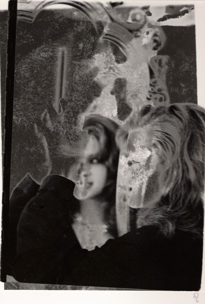The black-and-white image depicts two overlapping and distorted figures, with visible texture and marks giving it a worn or damaged appearance. One figure seems to be looking into a mirror, creating a reflection. There are decorative elements in the background, such as an ornate mirror frame. The scene has a surreal, abstract quality due to the degradation and layering of the image.