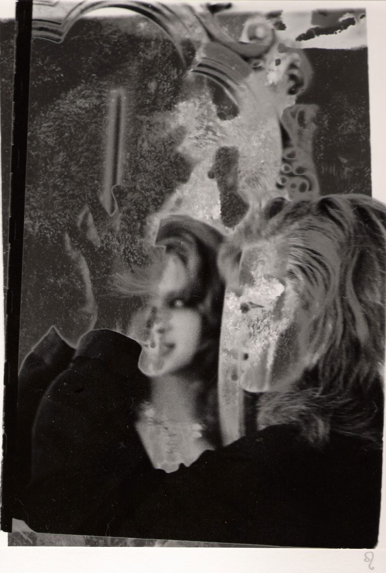 The black-and-white image depicts two overlapping and distorted figures, with visible texture and marks giving it a worn or damaged appearance. One figure seems to be looking into a mirror, creating a reflection. There are decorative elements in the background, such as an ornate mirror frame. The scene has a surreal, abstract quality due to the degradation and layering of the image.