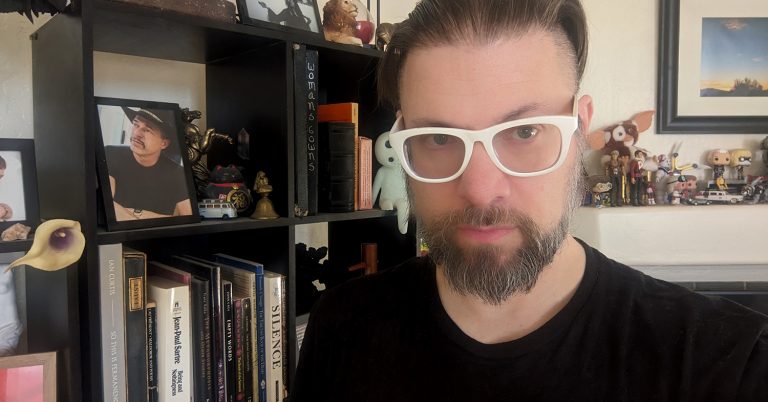 Me, with glasses and a beard standing indoors, in front of a black shelving unit filled with books, action figures, small sculptures, and knick-knacks. A framed photograph of my dad wearing a black shirt and hat is visible on the shelf. The words "Woman's Gowns" appear on a dark binder, and various collectible figures, such as a "Gizmo" from Gremlins and a small toy van, are displayed. A framed landscape photograph featuring a sunset is hung on the wall to the right.
