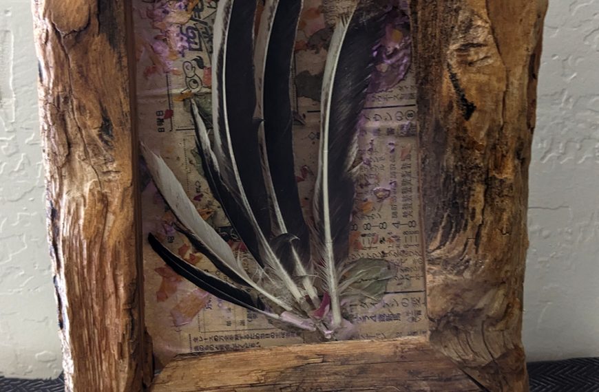 The image features a rustic wooden frame containing an arrangement of black and white feathers. The background inside the frame shows a collage of colorful, patterned paper. The frame is displayed on a black stand, set against a textured wall with a geometric patterned surface beneath.