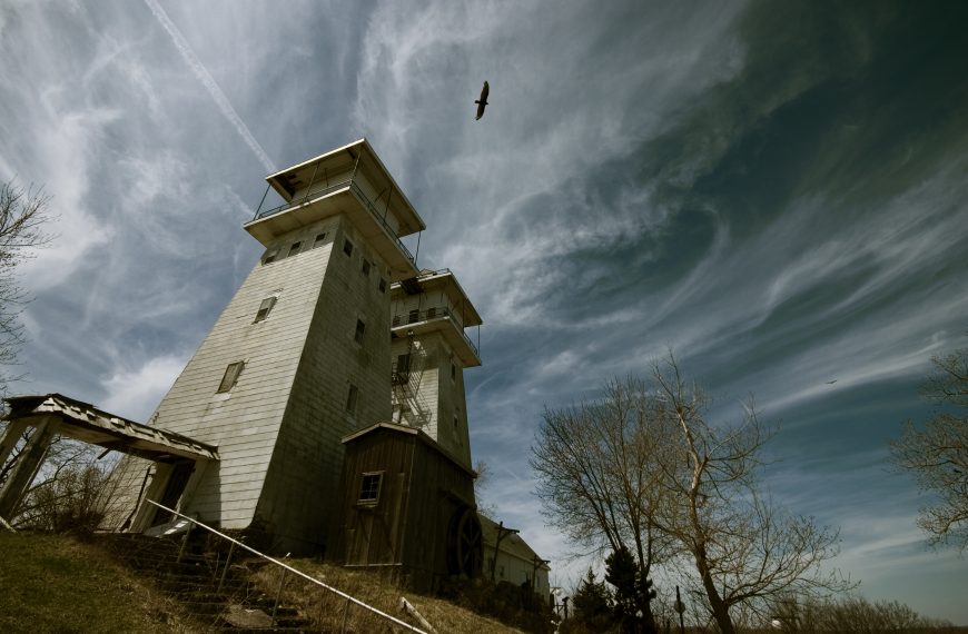 A low-angle shot captures a tall, weathered building with two prominent, shiplap-sided tower-like structures on a grassy hill under a dramatic, cloudy sky. An abandoned-looking wooden canopy extends from the side of the left tower, above steps with a metal railing. A small wooden shed and a large wooden wheel are visible at the base of the right tower. Bare-branched trees frame the scene, and a distinct airplane contrail cuts across the sky where a dark bird is in flight.
