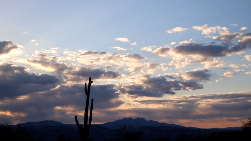 A desert landscape is depicted at sunrise or sunset, dominated by a vast sky filled with scattered clouds. The upper sky is light blue, transitioning to a mix of dark grey clouds illuminated by warm orange and yellow light peeking through from behind. In the foreground, a tall, silhouetted Saguaro cactus stands prominently. Behind it, dark mountain ranges stretch across the horizon, with other silhouetted desert vegetation visible at the very bottom.