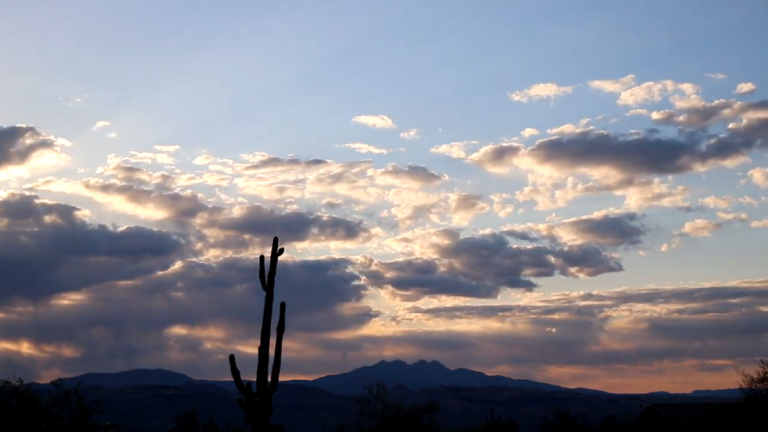 A desert landscape is depicted at sunrise or sunset, dominated by a vast sky filled with scattered clouds. The upper sky is light blue, transitioning to a mix of dark grey clouds illuminated by warm orange and yellow light peeking through from behind. In the foreground, a tall, silhouetted Saguaro cactus stands prominently. Behind it, dark mountain ranges stretch across the horizon, with other silhouetted desert vegetation visible at the very bottom.