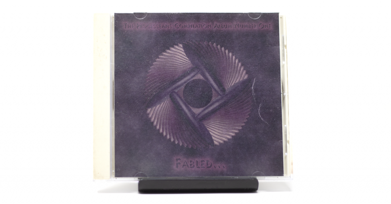 A CD in a clear plastic jewel case is positioned upright on a dark, slender stand against a plain white background. The CD cover features a dark purple and indigo background with a central abstract design. This design consists of a stylized, rotating motif with four ribbed, fan-like sections radiating from a dark central circle, forming a square outline. Text at the top reads "THE PROGRESSEANS COMPILATION ALBUM NUMBER ONE," and at the bottom, "FABLED..." is visible in a lighter shade.