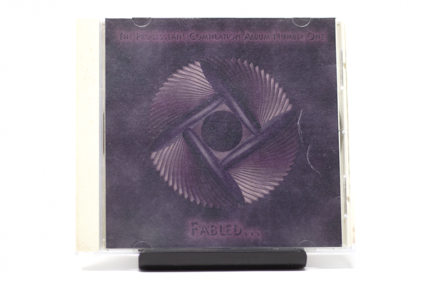 A CD in a clear plastic jewel case is positioned upright on a dark, slender stand against a plain white background. The CD cover features a dark purple and indigo background with a central abstract design. This design consists of a stylized, rotating motif with four ribbed, fan-like sections radiating from a dark central circle, forming a square outline. Text at the top reads "THE PROGRESSEANS COMPILATION ALBUM NUMBER ONE," and at the bottom, "FABLED..." is visible in a lighter shade.