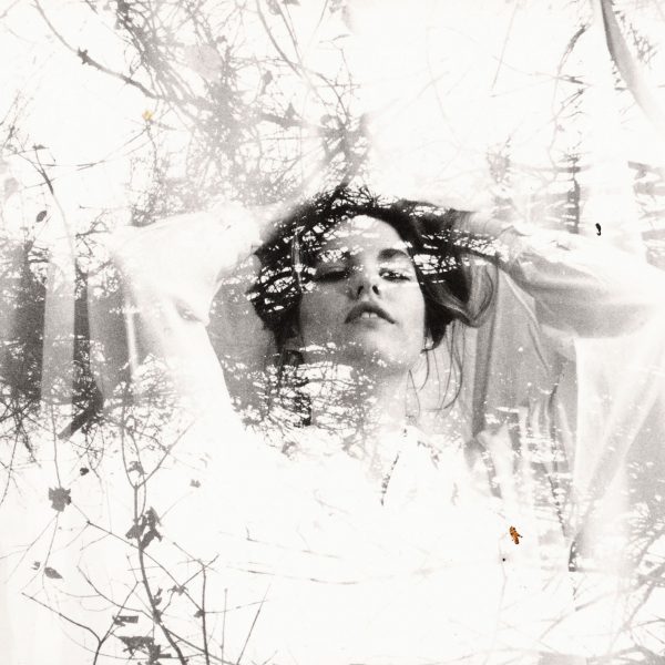 This is a black and white artistic double exposure photograph that blends the portrait of a person with natural imagery. The subject appears to be looking upward with a contemplative expression, their hair flowing naturally. Overlaid and integrated throughout the image are silhouettes of bare tree branches and twigs, creating an ethereal, dreamlike quality. The double exposure technique causes the natural elements to appear as if they're growing from or merging with the person's form, particularly around their head and upper body. The overall composition has a soft, ethereal quality with varying tones of gray and white, creating an artistic fusion between human form and nature.