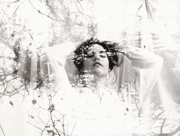This is a black and white artistic double exposure photograph that blends the portrait of a person with natural imagery. The subject appears to be looking upward with a contemplative expression, their hair flowing naturally. Overlaid and integrated throughout the image are silhouettes of bare tree branches and twigs, creating an ethereal, dreamlike quality. The double exposure technique causes the natural elements to appear as if they're growing from or merging with the person's form, particularly around their head and upper body. The overall composition has a soft, ethereal quality with varying tones of gray and white, creating an artistic fusion between human form and nature.
