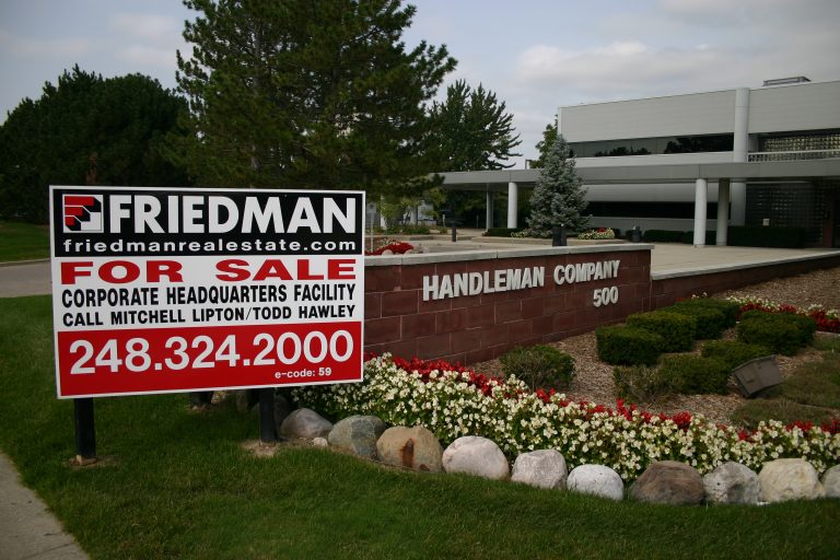 A wide outdoor shot captures the entrance to a corporate facility under an overcast sky. In the foreground on the left, a large red and black "Friedman Real Estate FOR SALE" sign advertises a "CORPORATE HEADQUARTERS FACILITY" with contact names "Mitchell Lipton/Todd Hawley" and phone number "248.324.2000". To the right of the sign, a reddish-brown brick-like monument prominently displays "HANDLEMAN COMPANY 500" in white letters. The base of the monument features landscaped beds with green bushes, rows of red and white flowers, and a line of light-colored rocks separating them from the green grass. In the background, a modern, light-colored corporate building with dark windows and a covered entrance supported by white columns is visible, partially obscured by various green trees, including tall pine trees and a smaller blue spruce.
