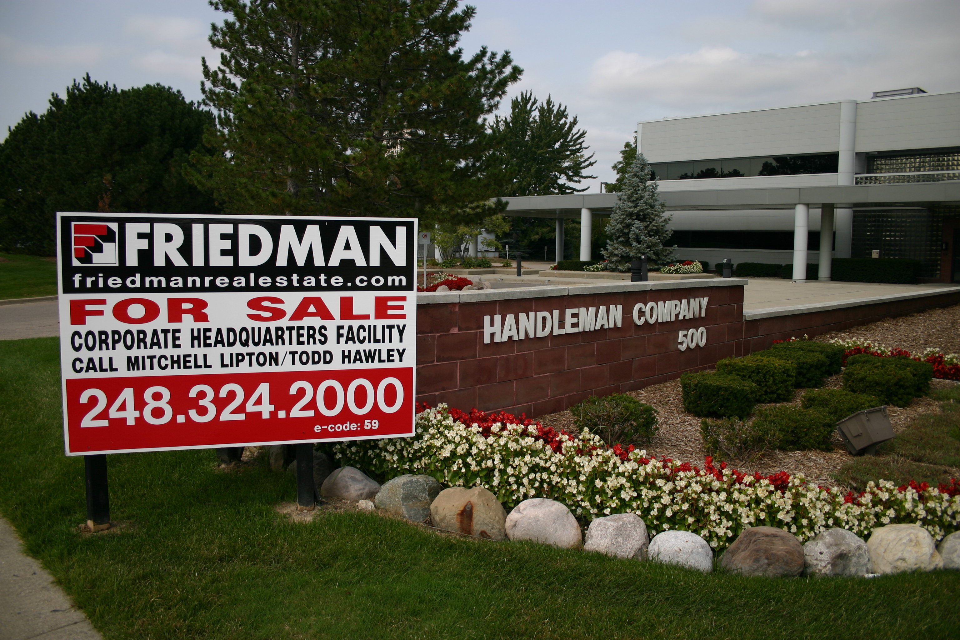 A wide outdoor shot captures the entrance to a corporate facility under an overcast sky. In the foreground on the left, a large red and black "Friedman Real Estate FOR SALE" sign advertises a "CORPORATE HEADQUARTERS FACILITY" with contact names "Mitchell Lipton/Todd Hawley" and phone number "248.324.2000". To the right of the sign, a reddish-brown brick-like monument prominently displays "HANDLEMAN COMPANY 500" in white letters. The base of the monument features landscaped beds with green bushes, rows of red and white flowers, and a line of light-colored rocks separating them from the green grass. In the background, a modern, light-colored corporate building with dark windows and a covered entrance supported by white columns is visible, partially obscured by various green trees, including tall pine trees and a smaller blue spruce.