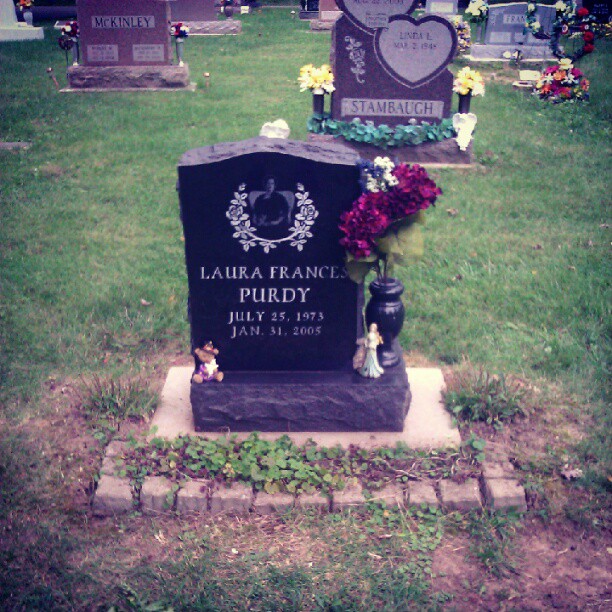 A black tombstone dedicated to "LAURA FRANCES PURDY" (JULY 25, 1973 - JAN. 31, 2005) stands in a grassy cemetery. The tombstone features an engraved image of a person within a white floral wreath, a dark vase holding purple and blue flowers, a small teddy bear, and a small angel figurine. In the background, other headstones are visible, including one marked "MCKINLEY" to the left and another heart-shaped stone to the right marked "LINDA L." and "STAMBAUGH".