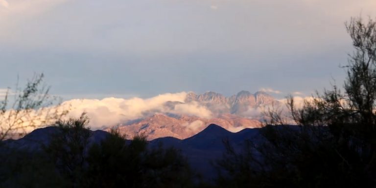 A panoramic landscape depicts multiple mountain ranges under a light blue and partly cloudy sky. Dark, out-of-focus brush occupies the foreground, framing midground mountains that transition from dark blue-purple to sunlit reddish-brown peaks. White and grey clouds drift over and between the higher mountain summits, illuminated by warm light.