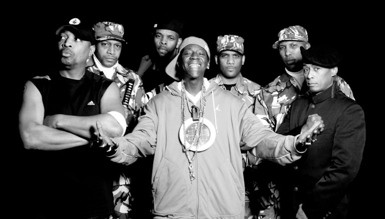 I can see a black and white photograph showing a group of seven men posing together. They're wearing a mix of casual streetwear and military-style camouflage clothing, including various caps and hats. The person in the center is wearing a light-colored hoodie with what appears to be a chain necklace and is smiling prominently. The group appears to be at some kind of event or gathering, with their arms around each other in a friendly group pose. The styling and aesthetic suggests this photo was taken in the 1990s hip-hop era.