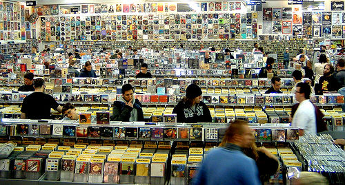 A bustling music store is filled with numerous people browsing through extensive rows of CDs and albums. The store's walls are completely covered with various album art, posters, and records, extending into the background. Many individuals, mostly young adults, are intently sifting through display bins filled with music, while others are standing and walking through the crowded aisles. A "DVD" sign is visible in the upper left, indicating a section for movies.