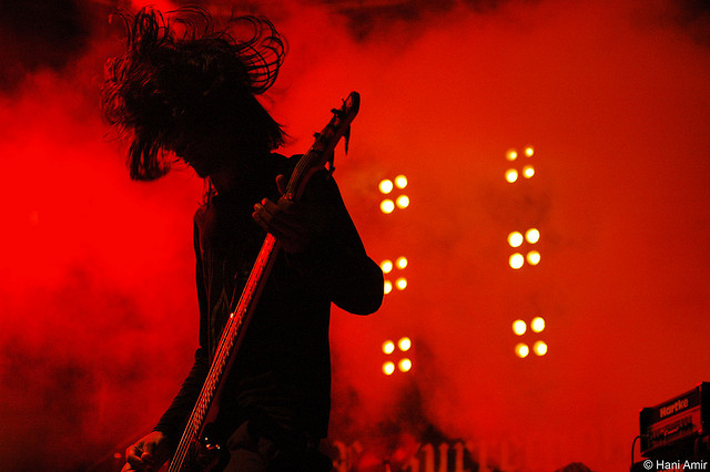 A heavily silhouetted musician is captured playing a bass guitar on a stage awash in red light and smoke. The musician's long hair is flung upwards, suggesting vigorous head movement. In the background, multiple sets of bright, circular stage lights illuminate the smoky atmosphere. A "HARTKE" brand amplifier is visible in the lower right portion of the image.
