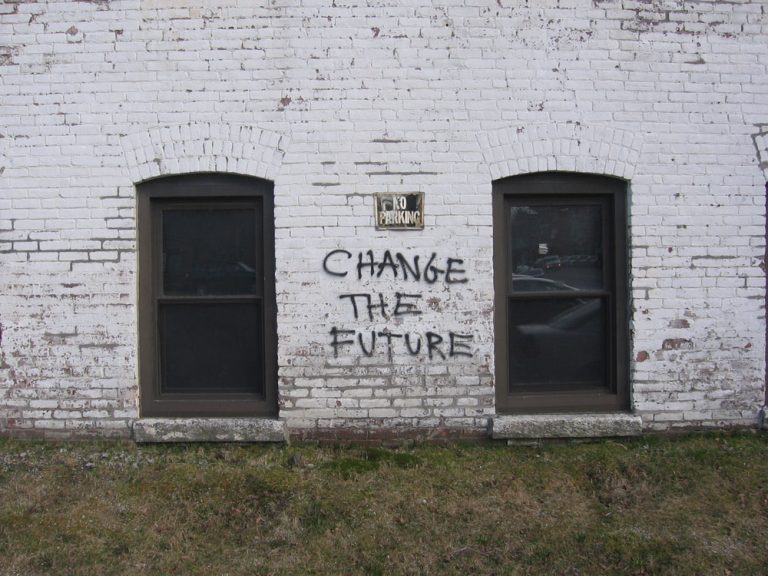 Graffiti reading "Change the Future" spray-painted in black on a weathered white brick building between two arched windows.
