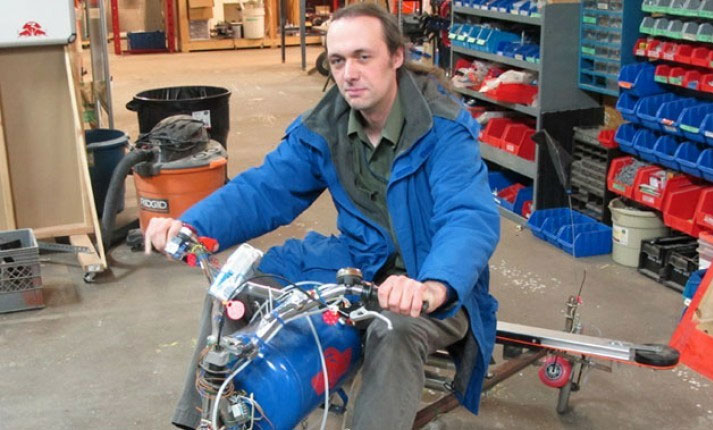 Man in a blue jacket sitting on a homemade or experimental motorized vehicle in a workshop.