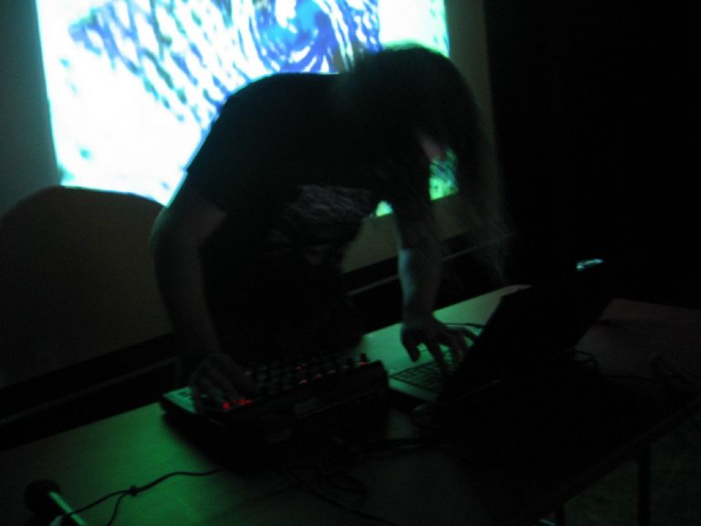 A person leaning over electronic music equipment and a laptop during a dark live performance, with abstract visuals