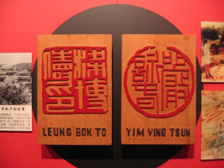 Two wooden carved Chinese seal plaques labeled "Leung Bok To" and "Yim Ving Tsun" mounted on a black circle against a red