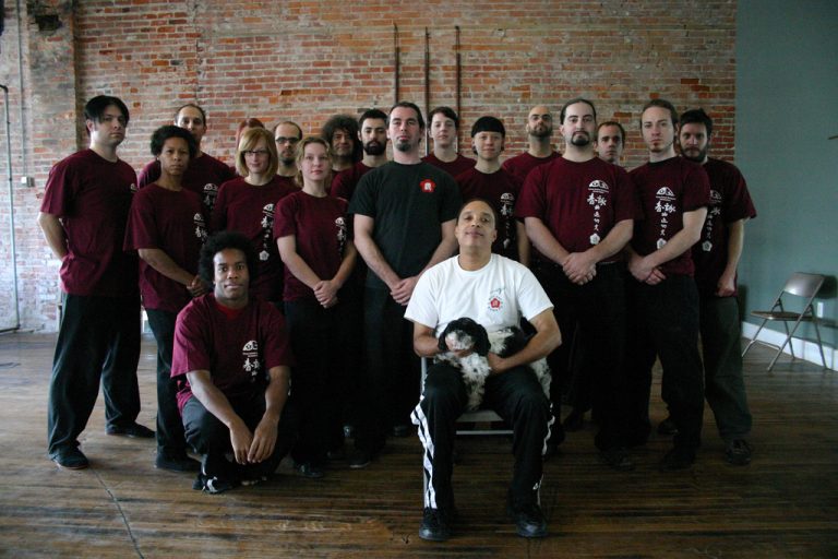 A martial arts group in matching dark red uniforms poses for a photo, with an instructor seated in front holding a black and