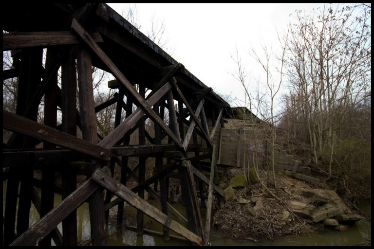 Deteriorating wooden railroad trestle bridge over a small creek, surrounded by bare winter trees.