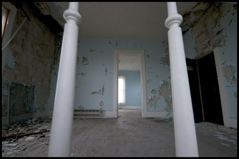Dilapidated room with peeling blue paint, debris-covered floor, two white bed posts in the foreground, and a doorway leading