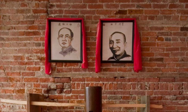 Two framed Chinese ink portrait drawings displayed on a red brick wall, each draped with red fabric.