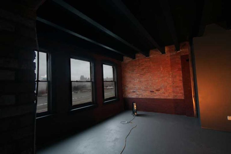 Empty industrial loft with exposed brick wall, dark ceiling beams, and three tall windows overlooking a city street.