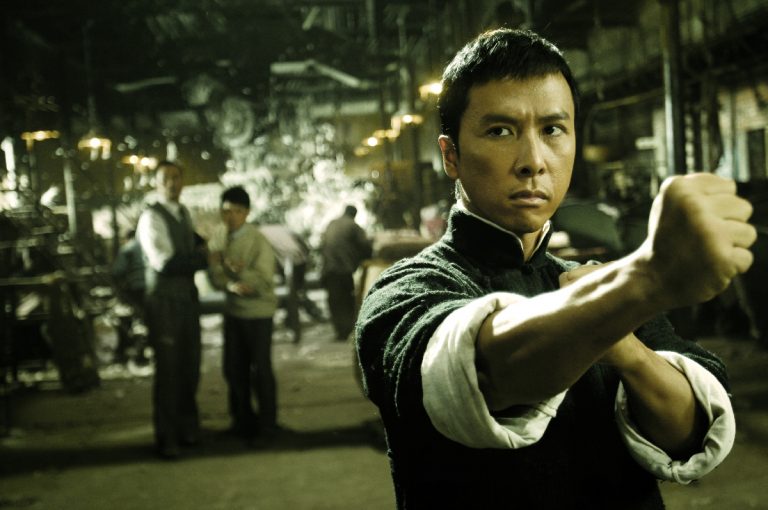 A martial artist in a black traditional Chinese jacket strikes a Wing Chun stance with fists crossed, inside a dimly lit
