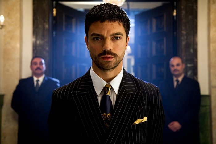 A well-dressed man in a pinstripe suit and tie stands confidently in a doorway, flanked by two men in dark suits in the